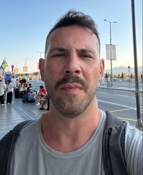 A man with a beard and gray t-shirt taking a selfie outdoors on a sunny day with a plaza in the background.