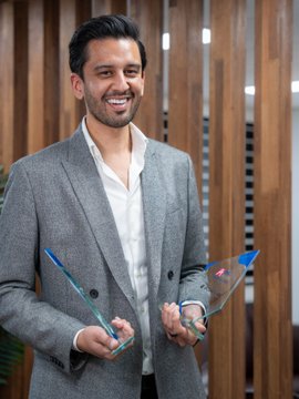 A professional headshot of a smiling man with dark hair and a beard, wearing a gray blazer over a white shirt and holding a pair of glasses, standing against a wooden slatted background.