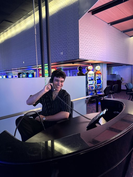 A young man with curly hair smiles while holding a telephone receiver at what appears to be a casino or gaming facility, with colorful slot machines visible in the background. The image pairs with his bio about his fintech background, suggesting a personal, casual moment as he reintroduces himself to his new audience.