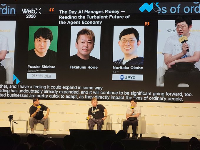 Three panelists—Yusuke Shidara, Takafumi Horie, and Noritaka Okabe—sit on stage at WebX 2026 discussing "The Day AI Manages Money – Reading the Turbulent Future of the Agent Economy," with JPYC branding prominently displayed alongside the discussion of AI-driven stablecoin adoption.