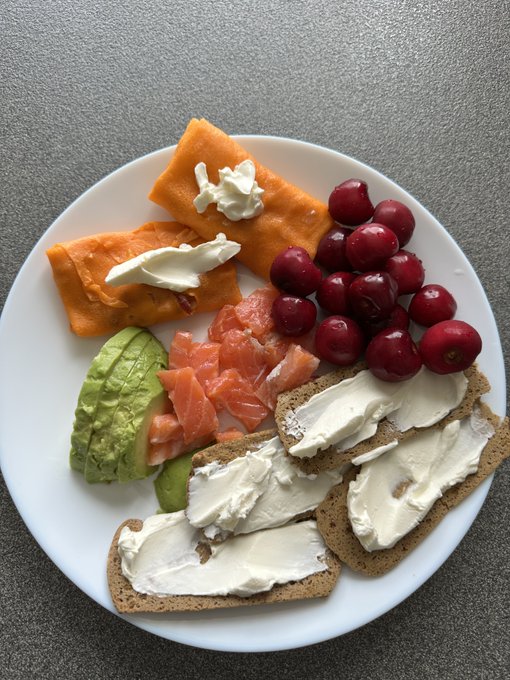 A white plate on a gray background holds a meal consisting of crackers with cream cheese, avocado slices, salmon, cherries, and orange slices topped with butter.