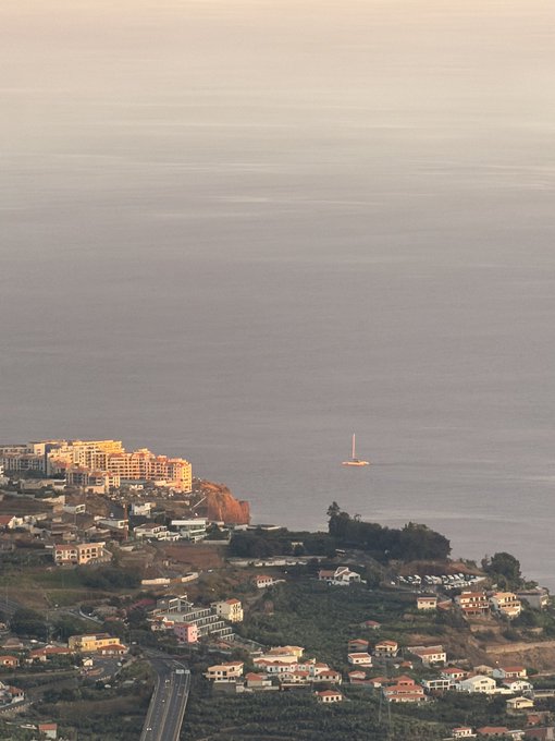 A coastal town stretches along a cliffside overlooking a calm sea at dusk, with a small sailboat visible in the water and a winding road cutting through the hillside neighborhood below. There is no text or signage visible in the image, and it appears unrelated to the accompanying post requesting a SOL (Solana) cryptocurrency address.