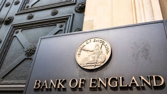 The image shows an ornate black door with decorative iron details and a sign bearing the "BANK OF ENGLAND" name alongside the bank's official seal. This imagery directly relates to the post about the UK government planning to expand the Bank of England's mandate to include supporting stablecoin and digital money innovation as a secondary objective.