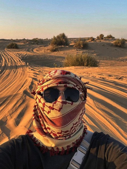 A person taking a selfie in the desert wearing a red and white keffiyeh wrapped around their head and face, along with aviator sunglasses.