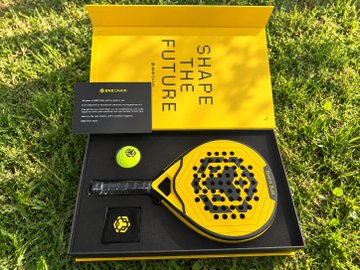 An open yellow box on grass displays a pickleball paddle and accessories with 'SHAPE THE FUTURE' and 'SELKIRK' branding on the packaging.