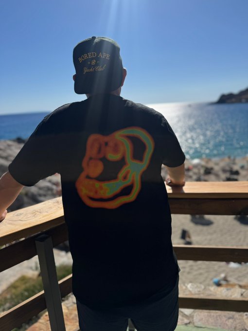 A person wearing a black "Bored Ape Yacht Club" t-shirt and matching cap looks out over a sunlit beach.