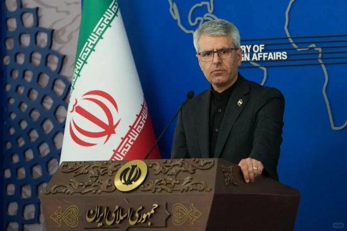 Iranian Foreign Ministry spokesperson Esmaeil Baghaei stands at a podium with the Iranian emblem, speaking in front of the Iranian flag and a blue backdrop reading 'MINISTRY OF FOREIGN AFFAIRS'.