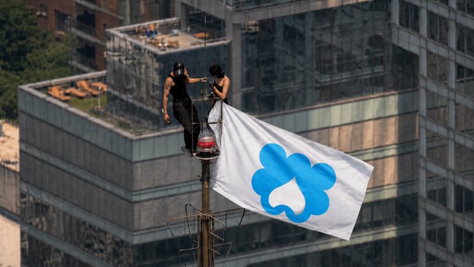 Two people stand on a tall pole or antenna structure high above city buildings, holding up a white flag with a blue cloud-like logo.