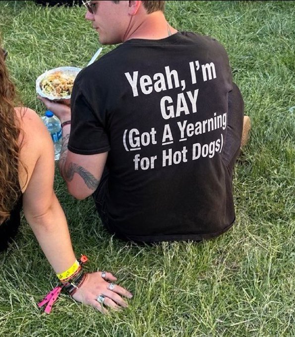 A person sitting on grass wears a black t-shirt with the text "Yeah, I'm GAY (Got A Yearning for Hot Dogs)" printed on the back.