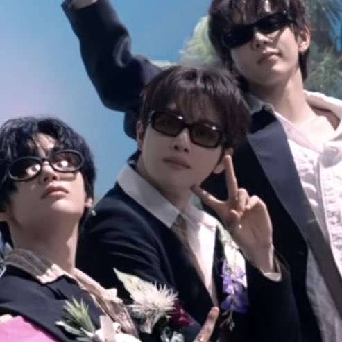 Three young men pose together outdoors wearing dark sunglasses and formal attire, with one making a peace sign gesture.