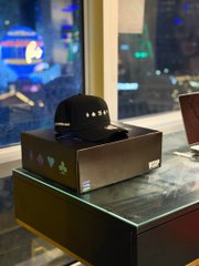 A black baseball cap rests on top of a dark green box on a marble surface, with a laptop partially visible and a cityscape in the background.