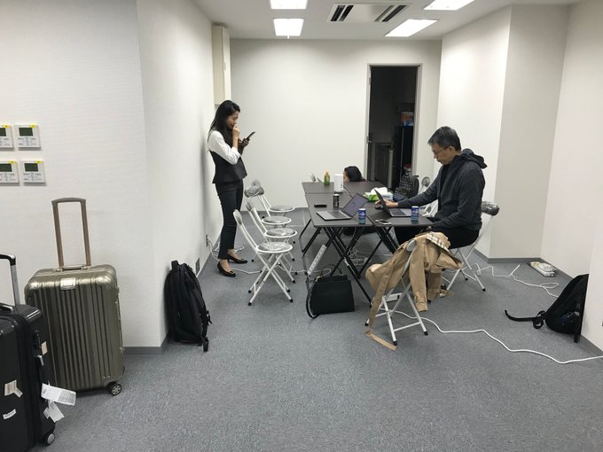 In this dimly lit room, several people are working on laptops around a large table, likely after arriving at a temporary office space as indicated by the luggage. The image shows a man at the table with laptops and drinks, while a woman stands on the side looking at her phone near her luggage.