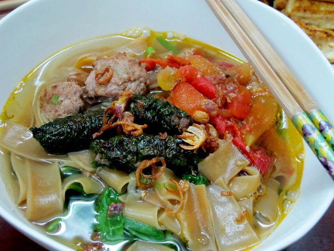 A bowl of Vietnamese soup featuring wide rice noodles, fried shallots, tomatoes, meat, and greens, served with chopsticks.