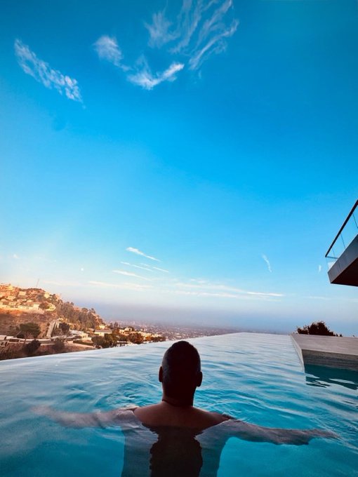 A man relaxes in an infinity pool overlooking a sprawling hillside city below a vast blue sky, embodying the ultimate "made it" moment after a 100x gain.
