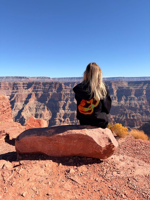 A person with long blonde hair sitting on a red rock at the Grand Canyon, wearing a black jacket with a colorful design, overlooking the vast canyon landscape under a clear blue sky.