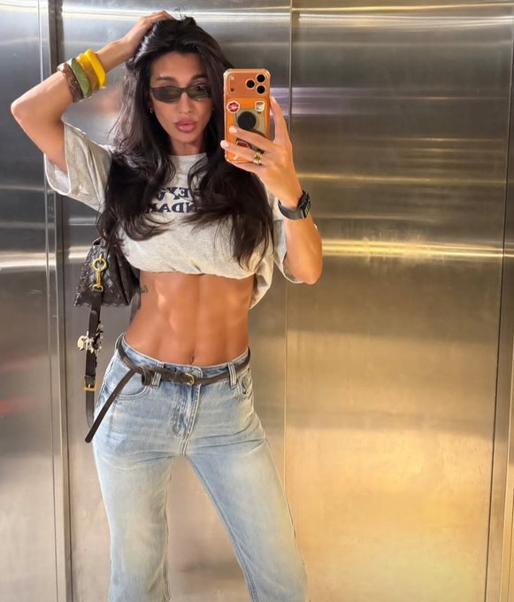 A woman takes a mirror selfie in an elevator wearing a cropped gray tee, low-rise light-wash jeans with a black belt, sunglasses, and a designer handbag, capturing a casual Friday vibe for the #crypto post.