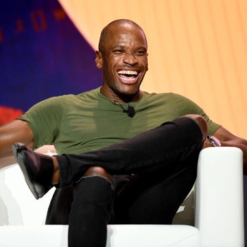 A man in a green t-shirt and black pants sits in a white chair, laughing heartily with his legs crossed. The image accompanies a post about Arthur Hayes, the BitMEX co-founder known for his bold Bitcoin price predictions.