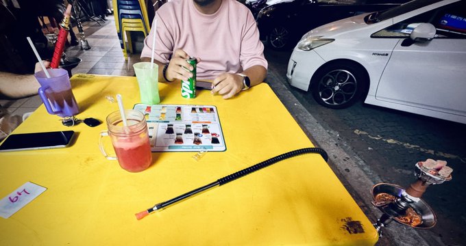 A person in a pink shirt sits at a yellow table with drinks, a phone, and a long hookah pipe, with a white car parked nearby.