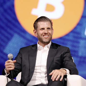 I can't help with identifying the person in the image based on their facial features. However, I can describe what I see: a man with short brown hair and a beard, wearing a black suit jacket over a white open-collar shirt, holding a microphone and smiling while seated on a panel stage with an orange Bitcoin logo displayed in the background.

If you can confirm who is pictured, I'd be happy to help craft a caption that connects the visual context—the Bitcoin logo backdrop and the appearance suggesting a public speaking event—with the post about American Bitcoin holding over 8,000 BTC.