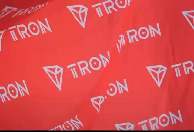 The image features a close-up view of a red fabric with white triangles on it. The text "TRON" can be seen written across the fabric, indicating that this might be related to the cryptocurrency or blockchain industry. The background appears to be blurred out, but there is some text visible in the bottom left corner which reads "The future of Web3 is being built by the next generation of innovators". This suggests a connection between the red fabric and the world of cryptocurrencies.