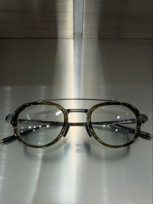 A pair of round, dark-rimmed eyeglasses with a double bridge rests on a reflective, brushed metal surface.