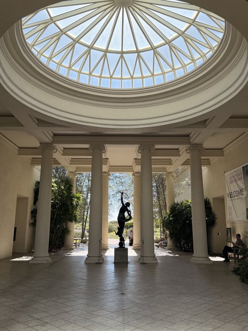 An interior view of a circular domed building with classical columns, a central statue, and potted plants visible through archways leading to an outdoor area.