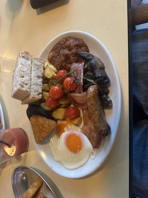 A full English breakfast on a white plate featuring baked beans, sausages, bacon, fried egg, mushrooms, cherry tomatoes on the vine, and bread, served on a yellow table.