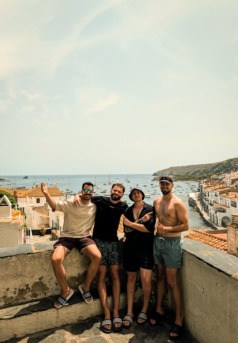 Four friends pose together on a stone wall overlooking a Mediterranean coastal village with boats in the harbor, embodying the camaraderie of "Mi equipo. Mis amigos. Mis hermanos." with relaxed smiles and arms around each other under a clear sky.