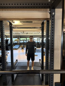 A man in black athletic wear stands inside a power rack in a gym, looking down at his phone.