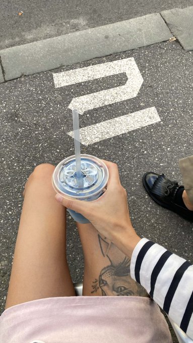 A first-person view of someone sitting outdoors holding a blue tumbler cup with a straw, with a tattooed leg visible and a painted "2" on the asphalt ground.