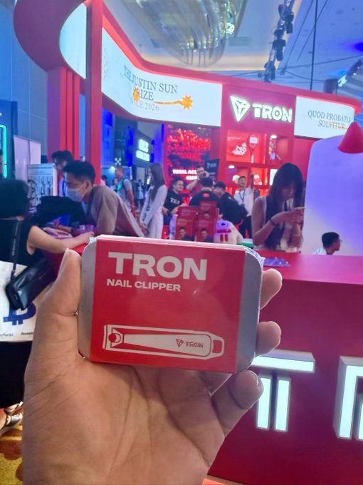 A hand holding a red and white promotional box labeled "TRON NAIL CLIPPER" at what appears to be a Justin Sun TRON booth event with crowds in the background.