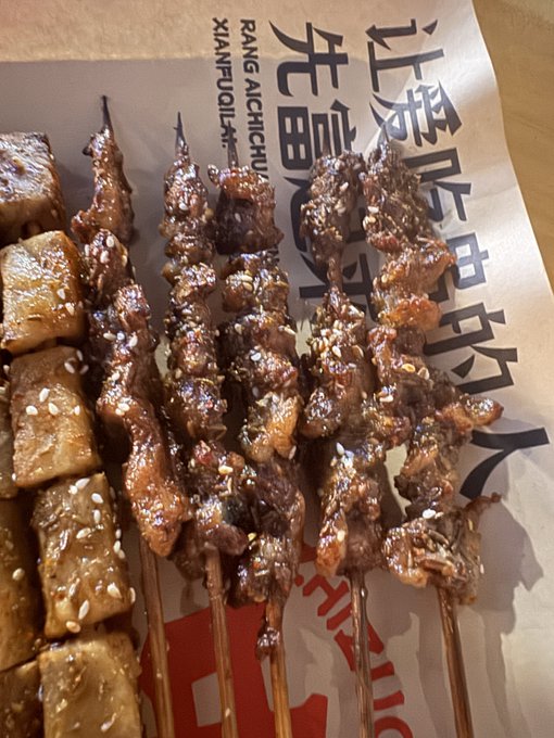 Skewers of grilled meat and tofu cubes sprinkled with sesame seeds resting on brown paper printed with 'PANG AICHCUNCHU XIANFULAKU' and Chinese characters.
