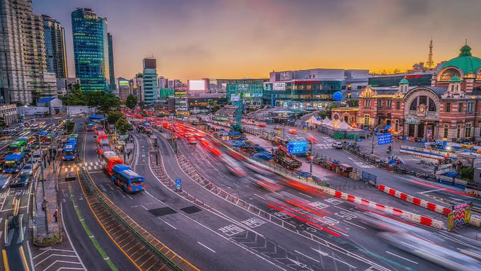 A vibrant cityscape at sunset shows a busy urban intersection with light trails from traffic, modern skyscrapers on the left, and a historic red-brick building with a green dome on the right.