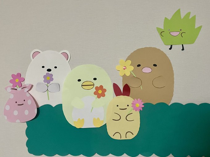 A craft project featuring cute paper cutout characters—a pink mushroom, white bear, green bird, yellow bunny, brown potato, and green leaf—arranged on a green scalloped base, holding small paper flowers.
