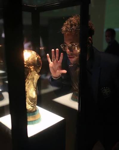 A person with glasses reaching out toward the FIFA World Cup trophy displayed inside a glass case.