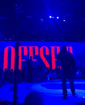 A performer stands on a stage bathed in blue lighting, with a large screen in the background displaying the red text "OBSESED."