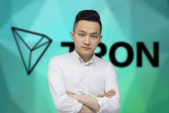 The image features a man standing with his arms crossed, wearing a white shirt. The background of the photo has a blue color scheme with a green triangle shape prominently displayed behind him. The text "TRON" can be seen in the image as well.