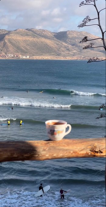 A coffee cup in the foreground with surfers and waves in the ocean behind it, set against a backdrop of mountains.