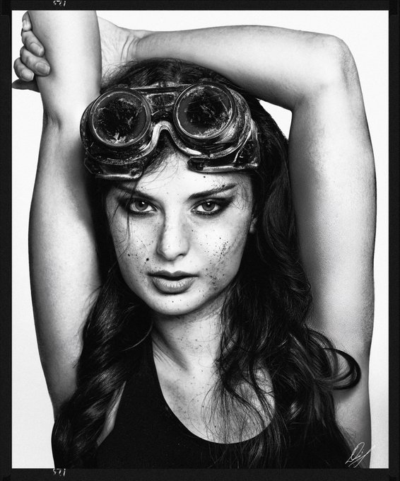 A black and white portrait photograph of a woman wearing large vintage goggles pushed up on her head, posing with her arms raised.