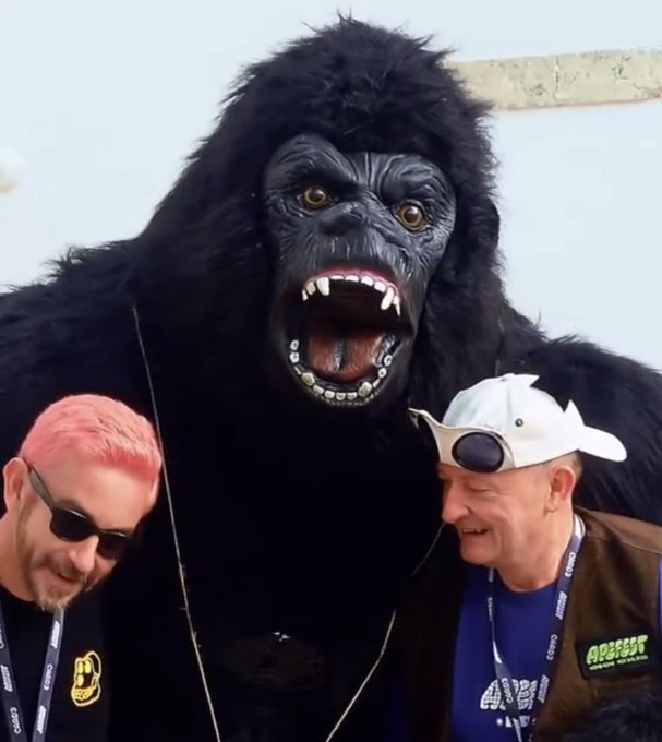 Two men pose with a person in a large gorilla costume that features an exaggerated mouth with fangs and grills.