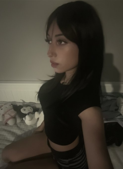 A young woman with long dark hair poses in a dimly lit room, wearing a black crop top with striped bottoms, while stuffed animals sit on a bed behind her. The moody, intimate aesthetic of the photo aligns with the cryptic, mysterious tone of the "encrypted" caption.