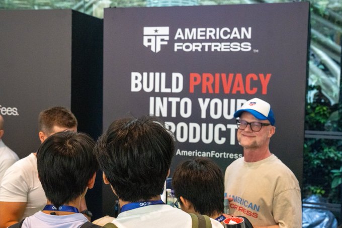A person wearing an American Fortress cap and glasses at a convention booth displaying 'BUILD PRIVACY INTO YOUR PRODUCT' with the AmericanFortress logo.