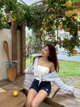 A young woman with long dark hair sits smiling on a wooden bench under a lemon tree arbor, wearing a white tube top with a light blue shirt tied around her shoulders and dark shorts.
