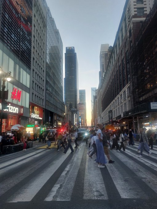 A street-level photo of pedestrians crossing a busy intersection in New York City, likely near Herald Square, with H&M and Verizon storefronts visible against a sunset sky.