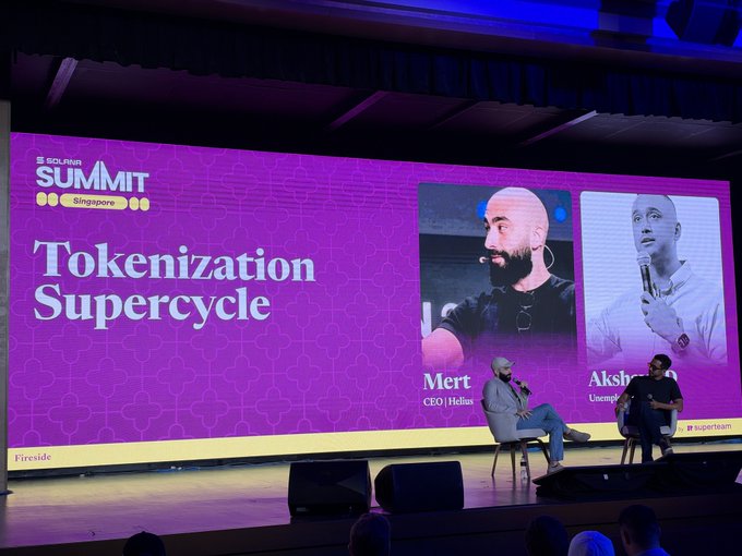 A fireside chat at Solana Summit Singapore titled "Tokenization Supercycle," featuring Mert (CEO of Helius) and Aksh on stage, with large screens displaying their portraits and titles behind them. The image directly captures the conference setting that the post references, where Mert's talk on the tokenization supercycle inspired the analysis of the four key bets (issuer, broker, fee share, DEX) in the Solana ecosystem.