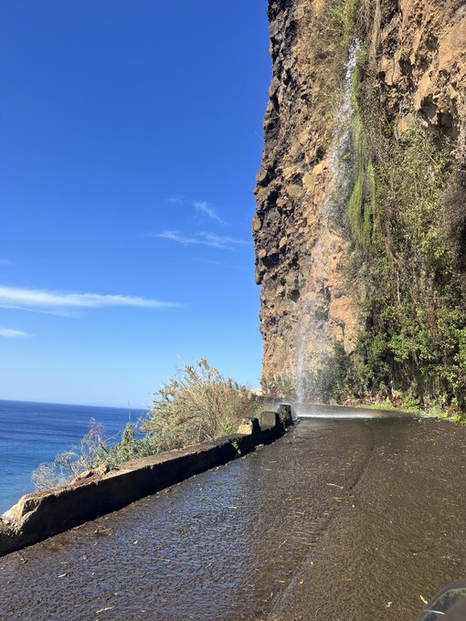 A scenic coastal road hugs a towering cliff with a waterfall cascading onto the wet pavement, overlooking the deep blue ocean under a clear sky—perfectly capturing the adventurous "Gm familia" vibe of starting the day with stunning tropical views.