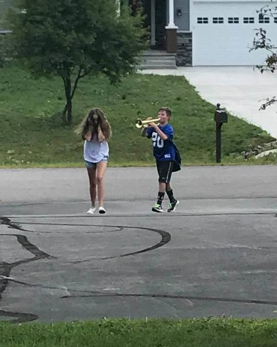 A boy in a blue #20 sports jersey playing a trumpet while walking down a residential street alongside a girl in a white t-shirt and denim shorts.