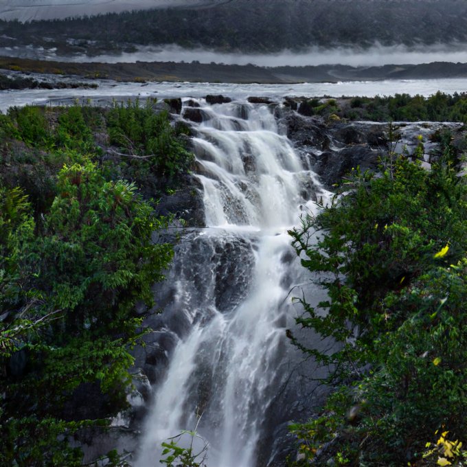 A waterfall cascading down a rocky cliff surrounded by green vegetation, with mist visible in the distant background.