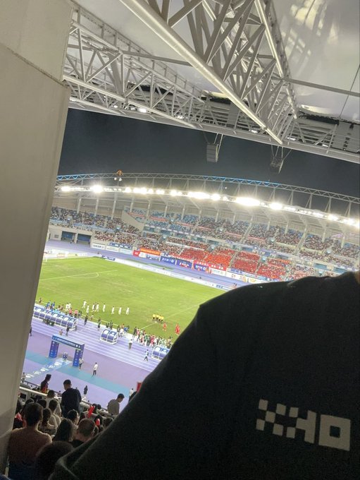A view from the stands of a large stadium at night, showing a soccer/football match in progress, with a person wearing a black hoodie with a pixelated logo sitting in the foreground.