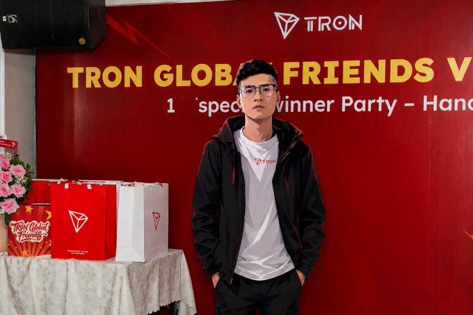 The image features a young man standing next to a red wall with a poster for "TRON GLOBAL FRIENDS VÔNG" written on it, which appears to be an event or gathering related to gaming. The man is wearing glasses and has his hands in his pockets. There are several bags of merchandise placed around the room, including some near him and others further away from him.